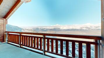 A balcony with a wooden railing overlooking a mountainous landscape with snow-capped peaks and a town below.