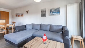 A modern living room with a grey sectional sofa, a wooden coffee table, and a small wooden stool. There are three framed pictures on the wall and a red flag on the coffee table. - Mélèzes R 020 - SKI LIFT apartment 8 people (Veysonnaz)