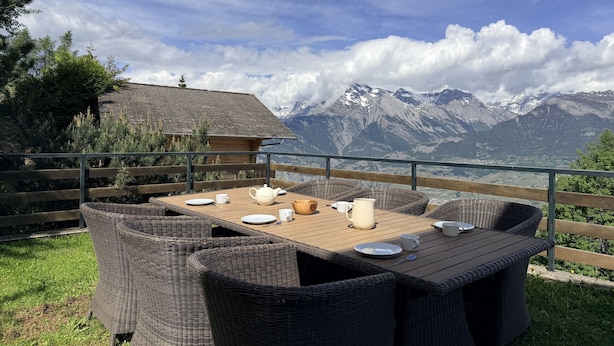 Une terrasse en bois, agrémentée de meubles en osier et d’une table dressée pour le repas, offrant une vue sur un paysage montagneux.
