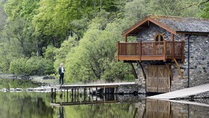 A wooden structure with a balcony, a stone wall, and a reflection in the water. - Luxury, romantic boathouse on the shores of Lake Ullswater, perfect for special occasions. (Ullswater)