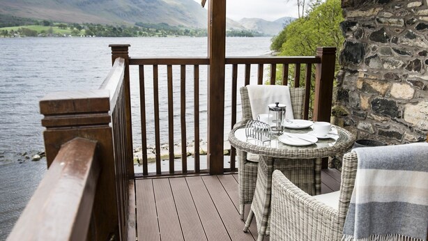 A wooden deck with wicker furniture overlooking a lake, stone walls, and a view of distant hills. - Luxury, romantic boathouse on the shores of Lake Ullswater, perfect for special occasions. (Ullswater)