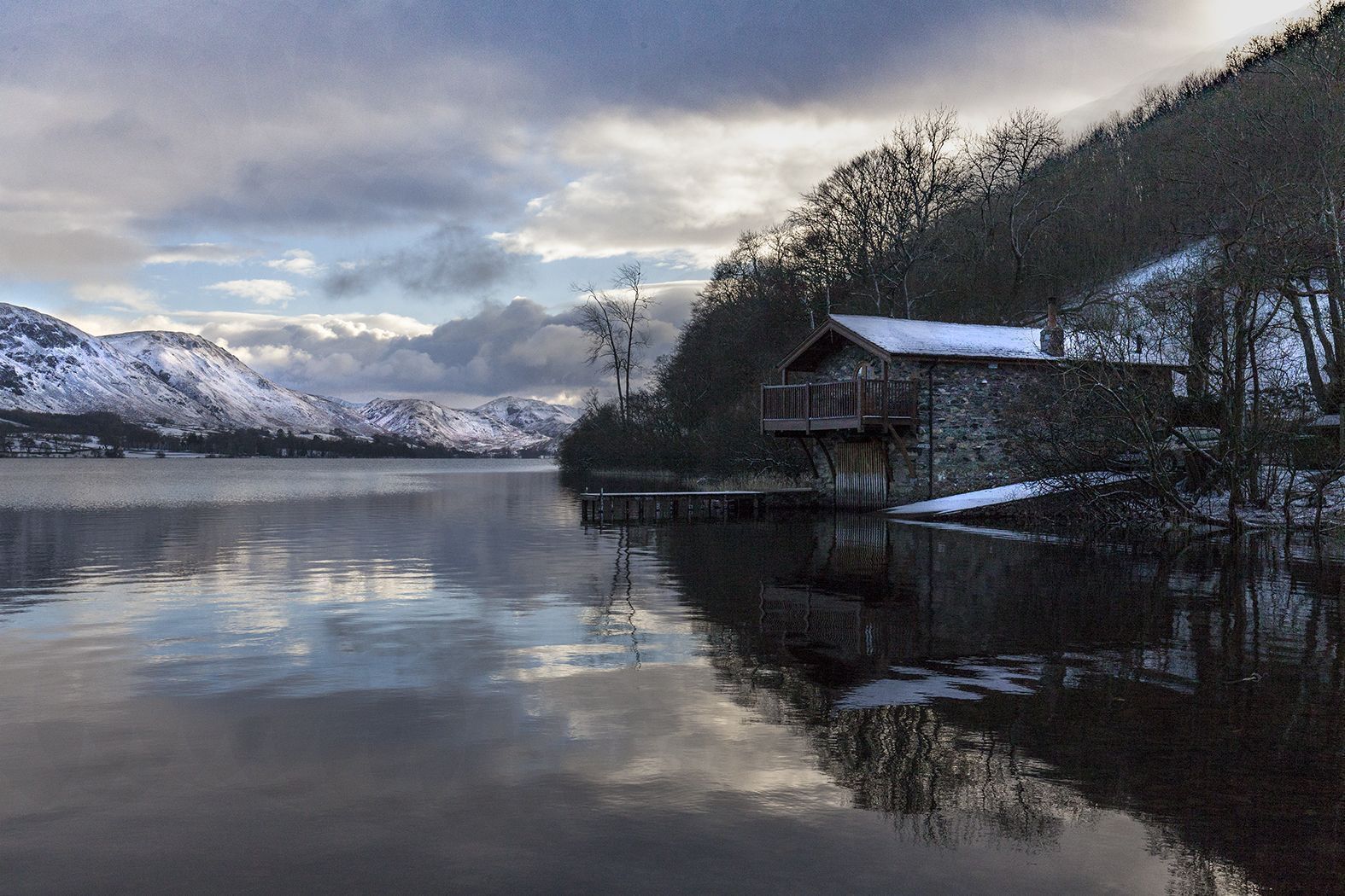 A wooden cabin with a balcony, situated by a calm lake with snow-covered mountains in the background.