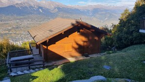 Eine Holzhütte mit Satteldach, einer Holztreppe, die zum Eingang führt, und einem Blick auf ein Tal sowie Berge im Hintergrund.