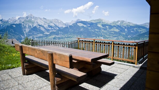 Ein Holztisch mit Bänken, vor einer Kulisse aus schneebedeckten Bergen und blauem Himmel.