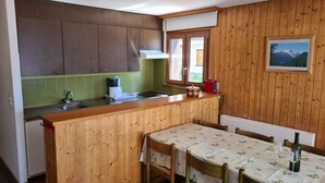 A compact kitchen with wooden cabinets, a green backsplash, a wooden dining table with a floral tablecloth, and a window with a mountain view. - Greppons P 003 - MOUNTAIN apartment 7 pers (Veysonnaz)