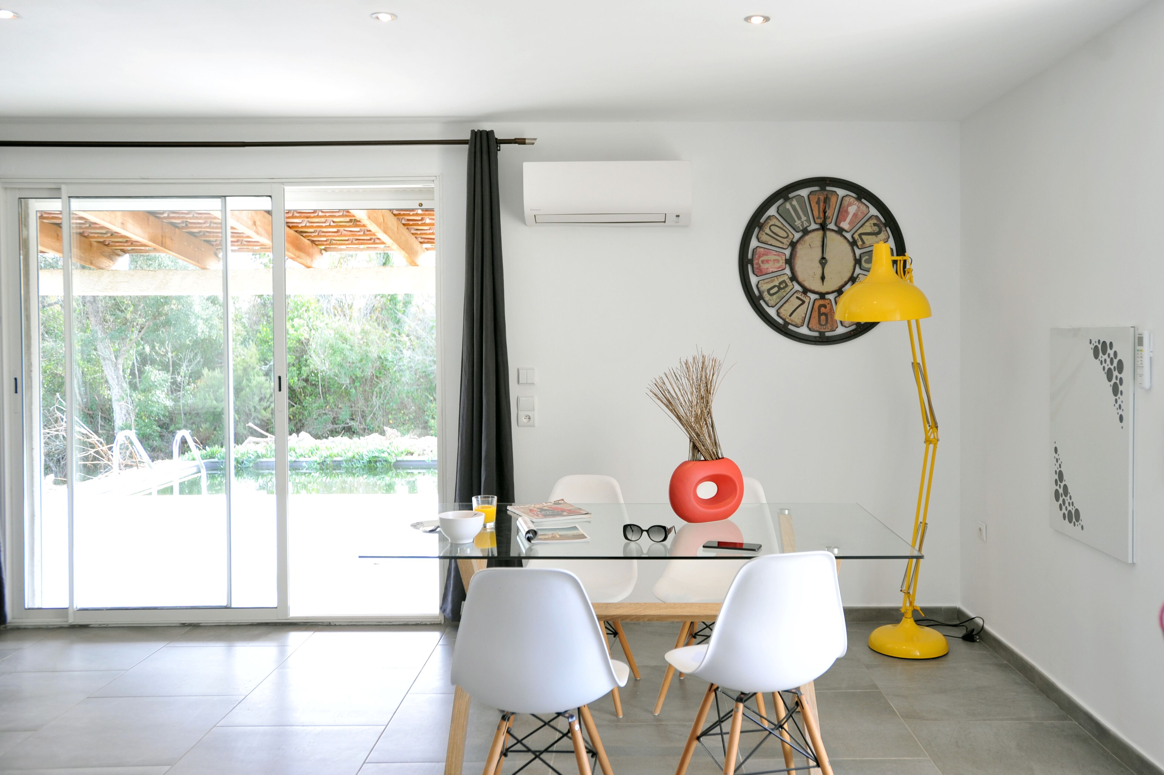 Un espace repas moderne avec une table en verre, des chaises blanches, un lampadaire jaune et une grande horloge murale.