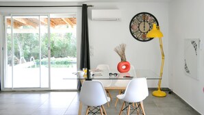 Un espace repas moderne avec une table en verre, des chaises blanches, un lampadaire jaune et une grande horloge murale.