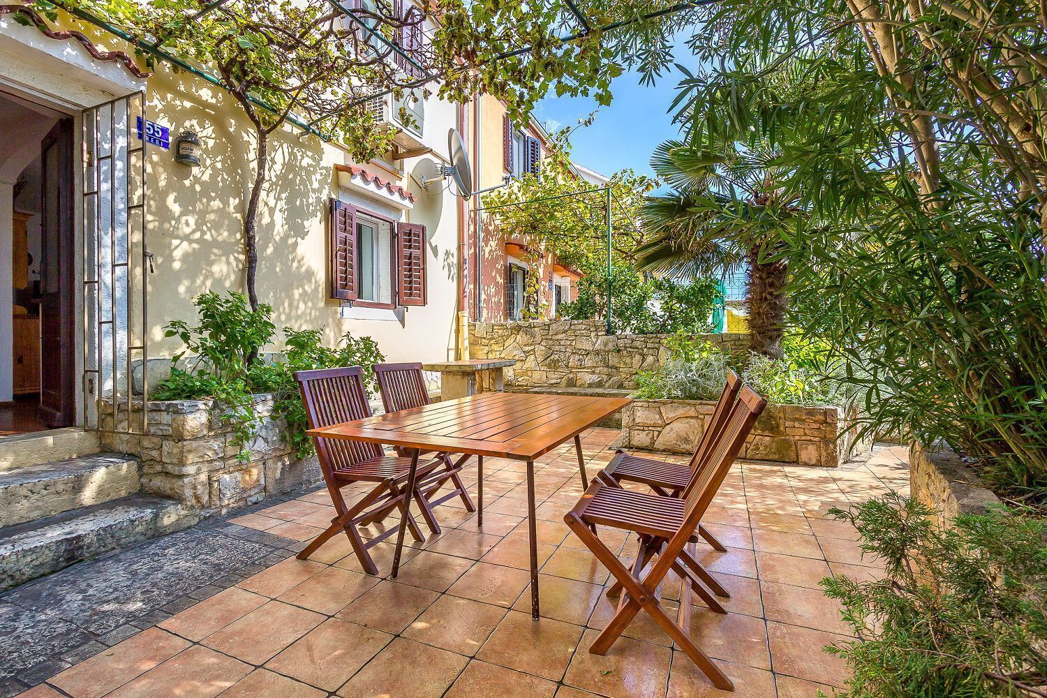 Un patio al aire libre con una mesa y sillas de madera, rodeado de muros de piedra y follaje.