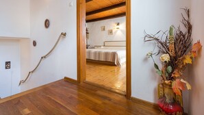 Um corredor com piso de madeira, uma cama ao fundo, um vaso decorativo com plantas secas e uma corda na parede.