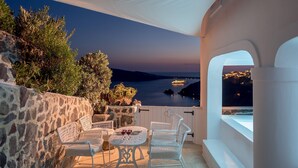Une terrasse dotée de meubles en osier blanc, une table avec des verres à vin et une vue sur la mer au coucher du soleil.