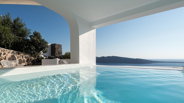 Une piscine à débordement à l’eau d’un bleu cristallin, ornée de colonnes blanches, et offrant une vue sur la mer et les collines lointaines.