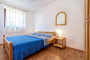 A single bed with a blue bedspread and matching pillows, a wooden nightstand with a lamp, a mirror on the wall, and a window with a floral curtain. - Large apartment with air conditioning, WiFi (Premantura)