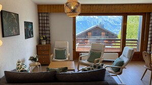 Un salon comprenant un canapé, deux fauteuils, une table basse en bois et une table d’appoint sur laquelle se trouve un vase. Deux tableaux ornent le mur, une fenêtre offre une vue sur un chalet en bois et le plafond est également en bois.