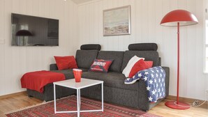 Uma sala de estar com um sofá cinzento, almofadas vermelhas, uma pequena mesa branca, um abajur vermelho e um tapete estampado.