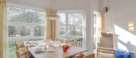 A dining area with a wooden table set for four, surrounded by chairs, large windows, and a view of trees.