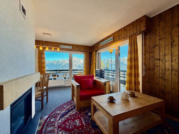 Una habitación con interior de madera, una chimenea, una mesa de comedor con una tetera, una silla roja y vistas a montañas a través de amplios ventanales.