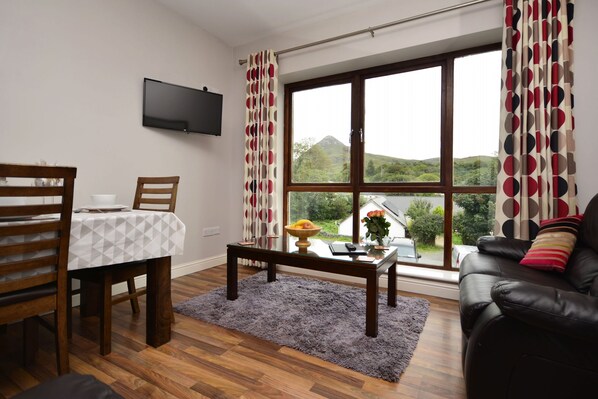 Ein Wohnzimmer mit Esstisch, Stühlen, einer Couch, einem an der Wand befestigten Fernseher und einem großen Fenster mit Vorhängen.