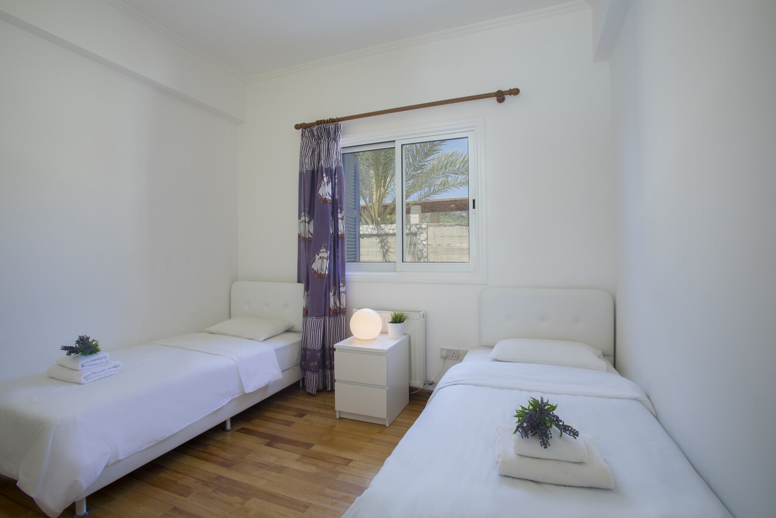 A room with two beds, a small table with a lamp, a window with a view of palm trees, and a curtain.