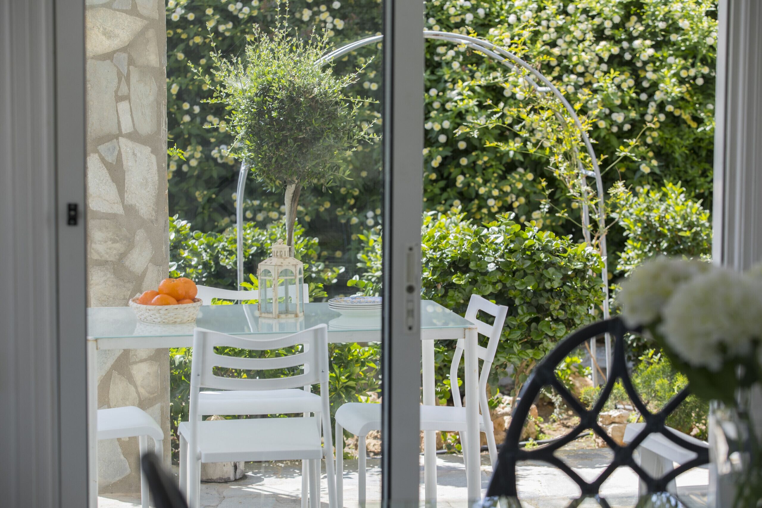 Une table en verre, des chaises blanches, un mur en pierre et un jardin avec différentes plantes et une cage à oiseaux.