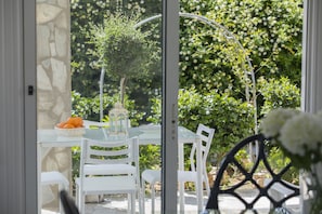 Une table en verre, des chaises blanches, un mur en pierre et un jardin avec différentes plantes et une cage à oiseaux.