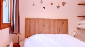 Un dormitorio con cabecera de madera, cama con ropa de cama blanca, lámpara de noche, ventana con cortina y estante con toallas dobladas.
