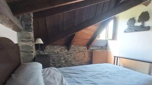 A bedroom with a stone wall, wooden ceiling beams, a bed with a patterned bedspread, a bedside table with a lamp, and a window with a view of trees. - The Balcony of Sotillo. Near Las Médulas (Sotillo de Cabrera)