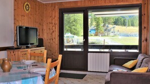 A room with a wooden interior, a dining table with chairs, a television, and a large window with a view of a park and buildings. - 2-Room Apartment, Sleeps 6, Ski-in/Ski-out, South Facing, Pets Allowed (Risoul)