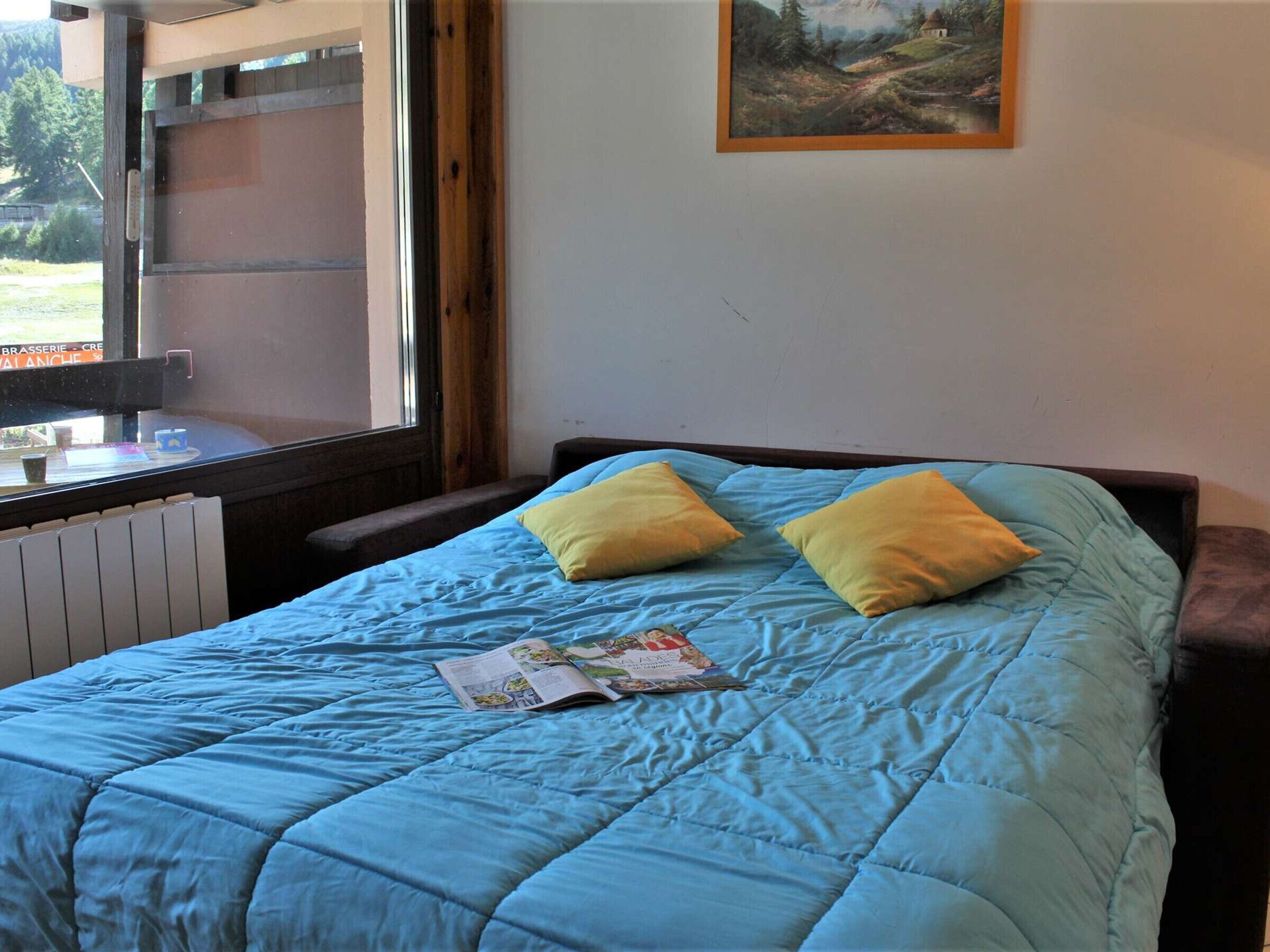 A bedroom with a blue bedspread, two pillows, a book, and a window with a view of a building and trees.
