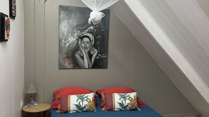 A bedroom with a sloped ceiling, a bed with red and blue bedding, a small bedside table with a lamp, and a wall-mounted painting. - Villa Marcaraïmon - between land and sea (Rivière-Pilote)