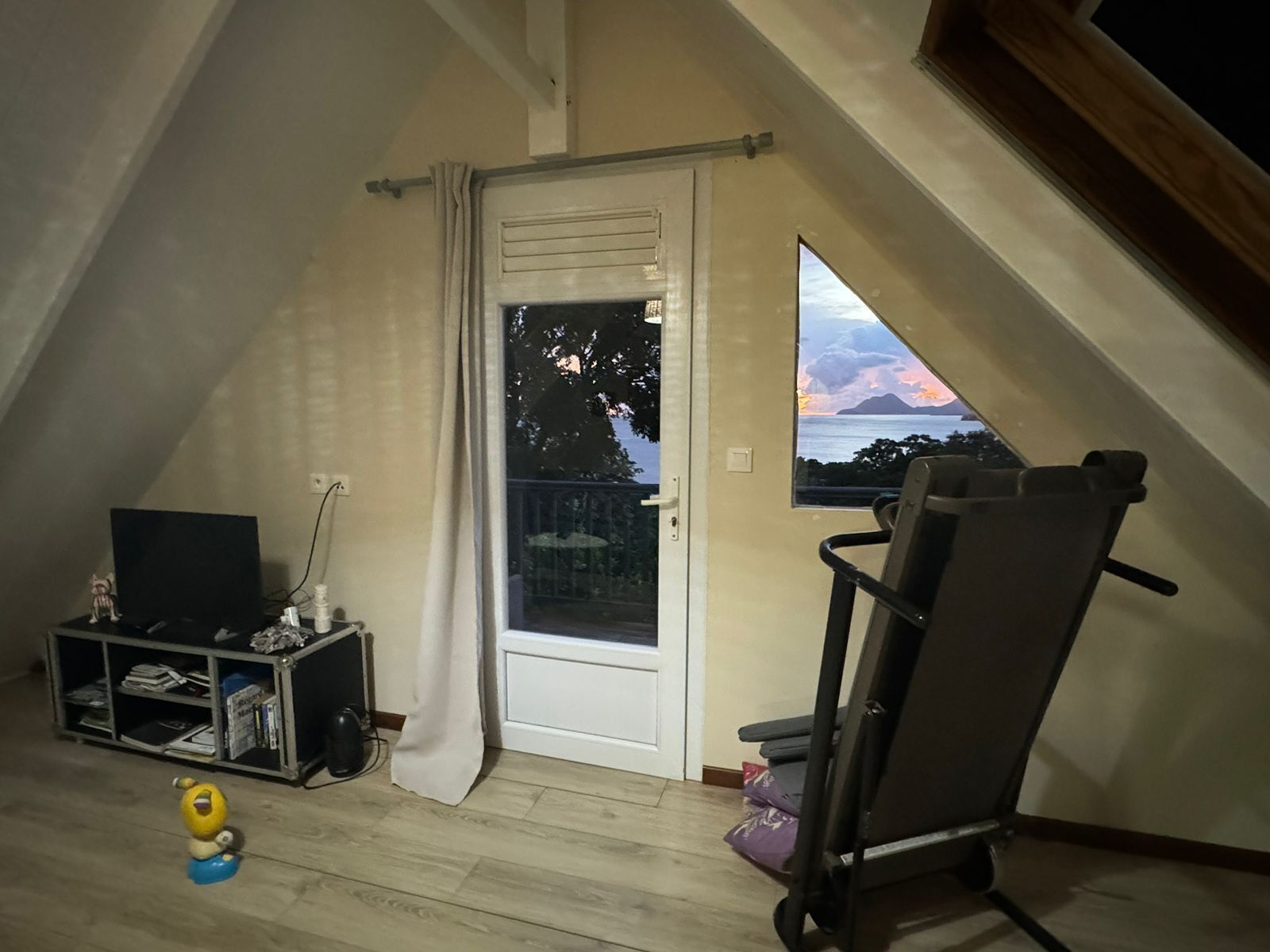 Een kamer met een schuine plafond, een glazen schuifdeur, een televisie, een klein plankje, een speelgoed en een stoel.