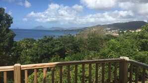 A coastal view with a wooden railing, dense greenery, and a distant town. - Villa Marcaraïmon - between land and sea (Rivière-Pilote)