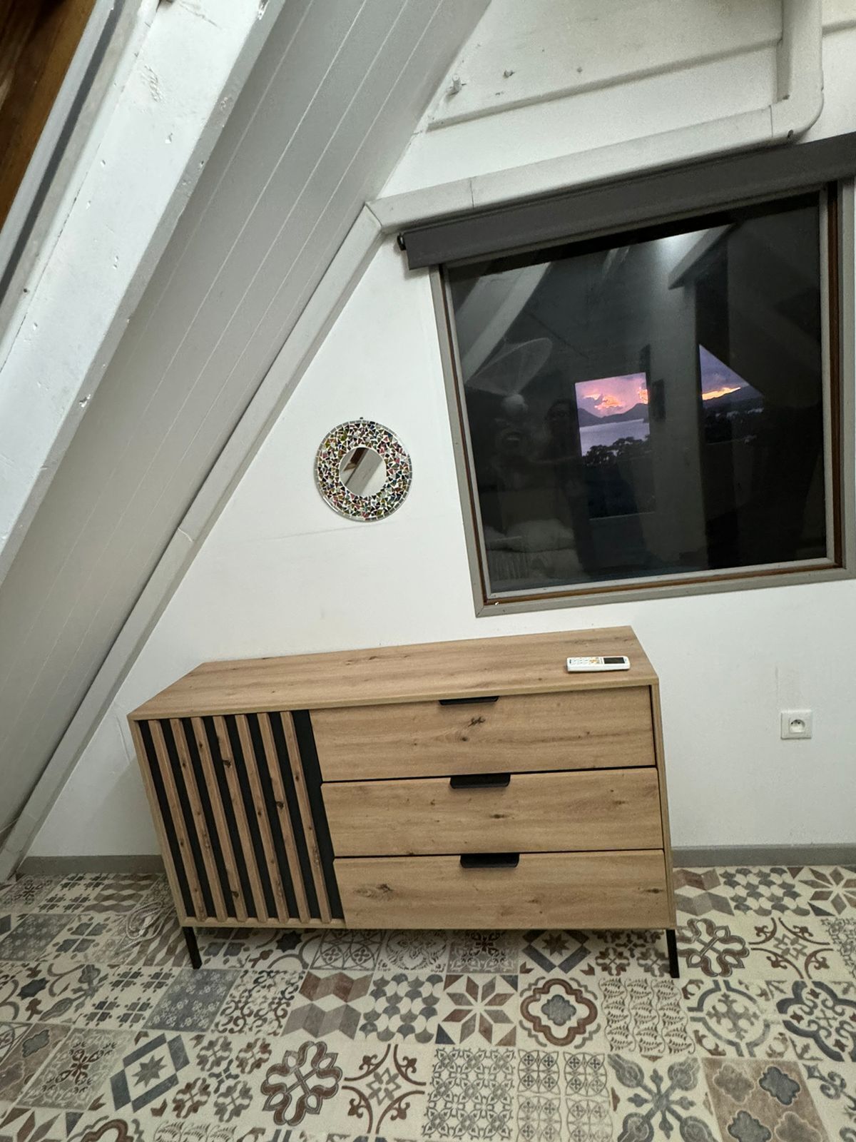 A room with a sloped ceiling, a window with a view of a sunset, a decorative mirror, and a wooden cabinet with drawers.