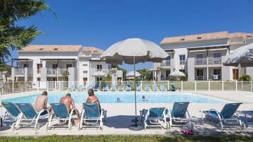 Zona de piscina con sillones y sombrillas, rodeada por un edificio con balcones.