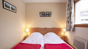 Een tweepersoonsbed met twee rode dekens, twee bedlampjes, een fotocollage aan de muur en een raam met gordijnen.