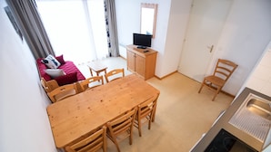 Ein gemütlicher Wohnbereich mit Esstisch aus Holz, vier Stühlen, einer kleinen Küchenzeile mit Herd, einem Fernseher auf einem Holzschrank und einer Couch mit Kissen.
