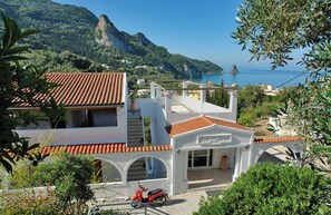 Un edificio costero con techo de tejas rojas, fachada blanca y vista al mar y a las montañas.