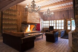A log cabin interior with a fireplace, leather seating, chandeliers, and a stone wall.