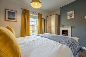 A bedroom with a bed, a window with curtains, a fireplace, and a painting on the wall. - Samphire Cottage (BS), Brancaster Staithe, Norfolk (King's Lynn)