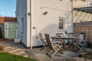 A patio area with a round table and four wooden chairs, a green door, and a white building with a window. - Samphire Cottage (BS), Brancaster Staithe, Norfolk (King's Lynn)