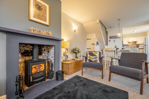 A cozy living room with a fireplace, a dining area, and a staircase leading to another level. - Samphire Cottage (BS), Brancaster Staithe, Norfolk (King's Lynn)