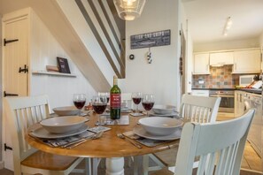 A dining area with a wooden table set for four, surrounded by white chairs. A bottle of wine and glasses are on the table. - Samphire Cottage (BS), Brancaster Staithe, Norfolk (King's Lynn)