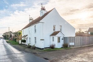 A white two-story house with a sloped roof, a front porch, and a small garden. - Samphire Cottage (BS), Brancaster Staithe, Norfolk (King's Lynn)