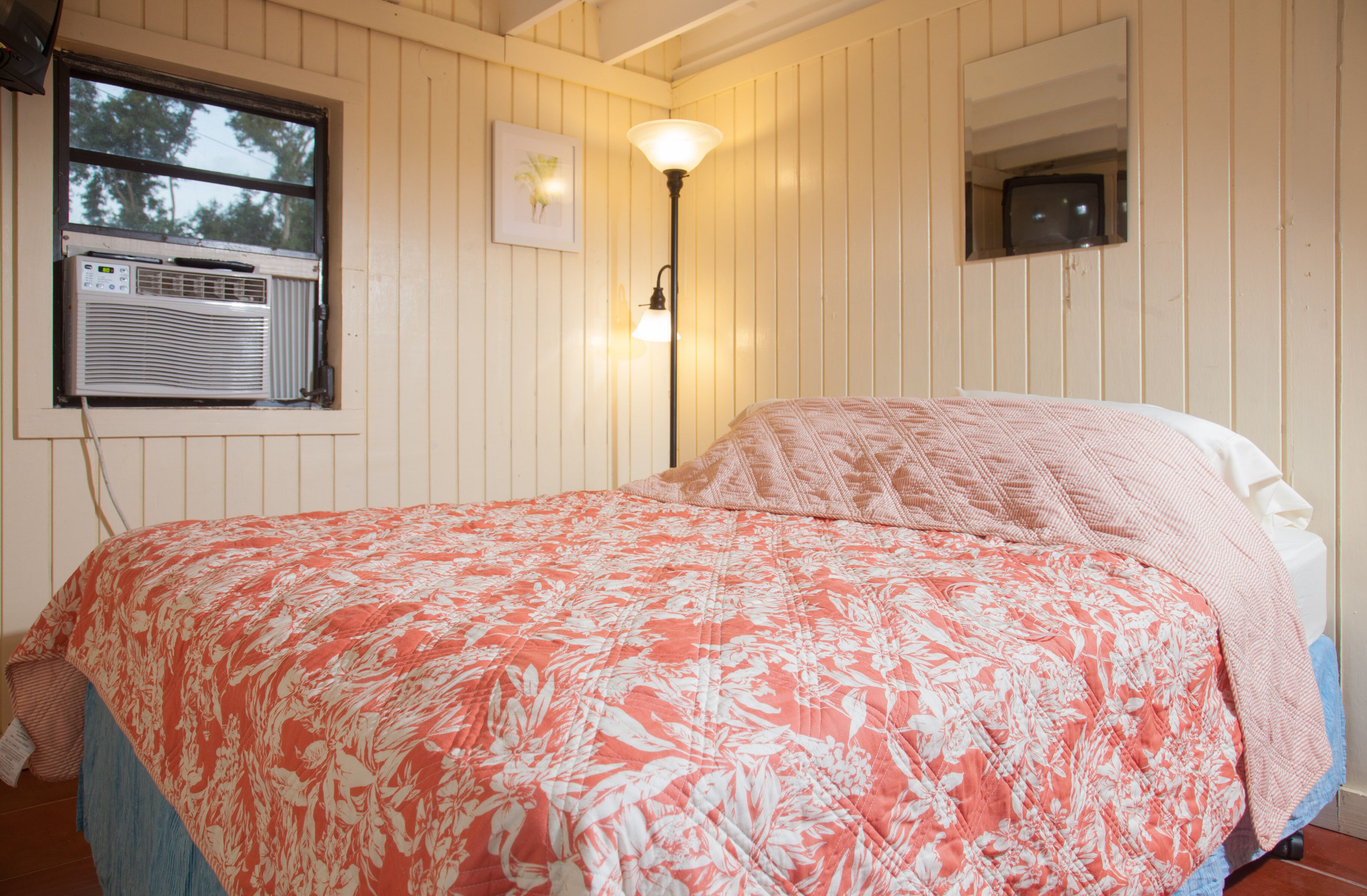 Une chambre à coucher avec un lit, une fenêtre équip ée d’un climatiseur, une lampe fixée au mur et un petit téléviseur.