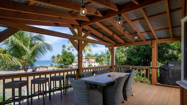 A covered outdoor deck with a wooden table and wicker chairs, overlooking a beach with palm trees and the ocean.