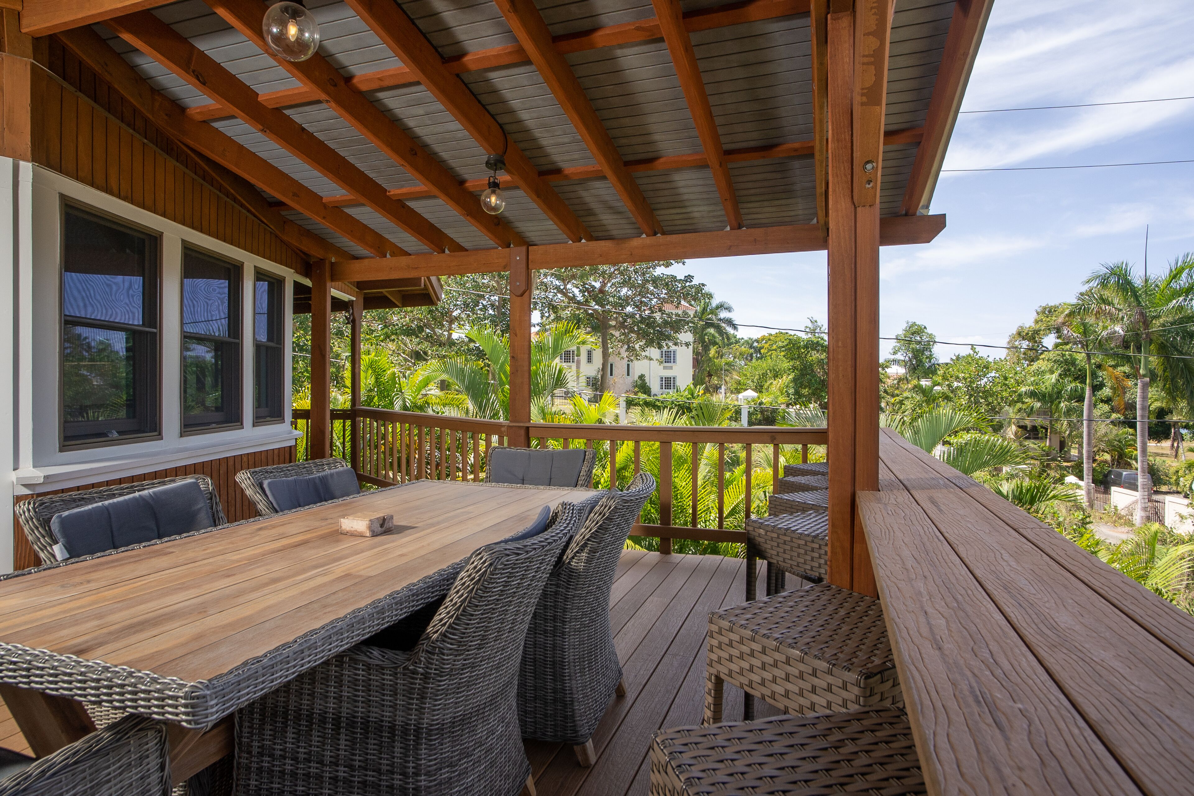 A covered outdoor deck with a wooden table and wicker chairs, overlooking a lush garden and a residential area.
