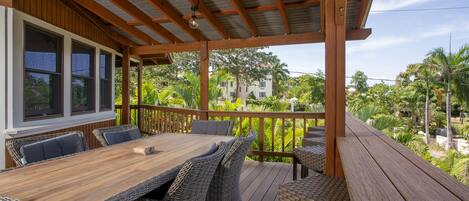 A covered outdoor deck with a wooden table and wicker chairs, overlooking a lush garden and a residential area.