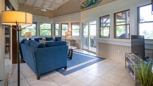 A spacious living room with a blue sofa, a ceiling fan, large windows, and a TV.