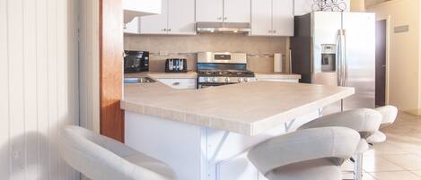Cocina moderna con armarios blancos, refrigerador de acero inoxidable, horno empotrado y una zona con taburetes de bar.