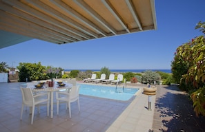 A pool area with a covered patio, lounge chairs, and a view of the sea.