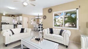 A spacious living room with two white sofas, a glass coffee table, and a ceiling fan. A kitchen with white cabinets and a dining area with a table and chairs are visible in the background. - PALM BEACH LUXURY VACATION  VILLA !!!!! (Palm beach)